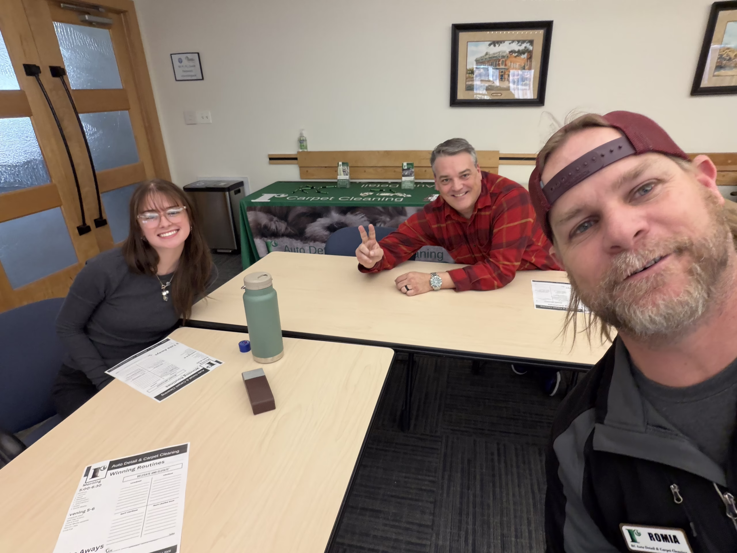 Three adults are sitting around a table in a classroom or meeting room, smiling at the camera. Two are seated with paperwork in front of them, and one is taking a selfie, wearing a cap backwards.