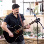 A man with dark hair and a mustache plays a black acoustic guitar and stands in front of a microphone indoors. There are plants and blurred people in the background.