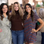 Three women smiling and posing together at an indoor event. One wears a striped dress, one has a brown top with jeans, and the third wears a striped short-sleeve dress. People and activity are visible in the blurred background.