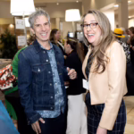 A man in a denim jacket and a woman in glasses and a tan blazer smile and laugh together at a busy indoor event with other people mingling in the background.