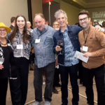 Five people stand together smiling at a networking event; one woman wears a yellow hat, others are dressed in business casual attire. A crowd mingles in the background under the "BizWest" sign.