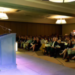 A woman stands at a podium speaking to a large seated audience in a conference room with high ceilings and hanging lights. The audience listens attentively under warm lighting.