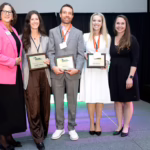 Five people stand on a stage smiling, three holding award plaques. One woman wears a bright pink blazer, another a white dress, and the rest are in business attire. A podium is visible to the left and a black curtain is in the background.