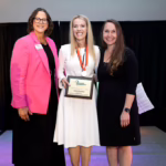 Three women stand together smiling at an indoor event. The woman in the center, wearing a white dress and a medal, holds a framed award. The woman on the left wears a bright pink blazer, and the woman on the right wears a black dress.