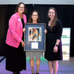 Three women stand on a stage, smiling and holding a framed award together. One woman wears a pink blazer, the others are in dresses. The background has black curtains and illuminated "Community" signage.