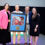 Four people stand on a stage smiling; one man holds a large framed artwork with a car illustration and plaque. Two women stand on either side of the group, one in a pink blazer and one in a black dress.