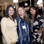 Three women pose together, smiling at an indoor event. They each wear name badges and patterned outfits. Other people mingle in the softly lit background. The atmosphere appears friendly and social.