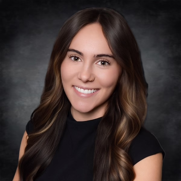 A woman with long, wavy brown hair and light highlights smiles at the camera. She is wearing a black top and is posed in front of a dark, softly blurred background.