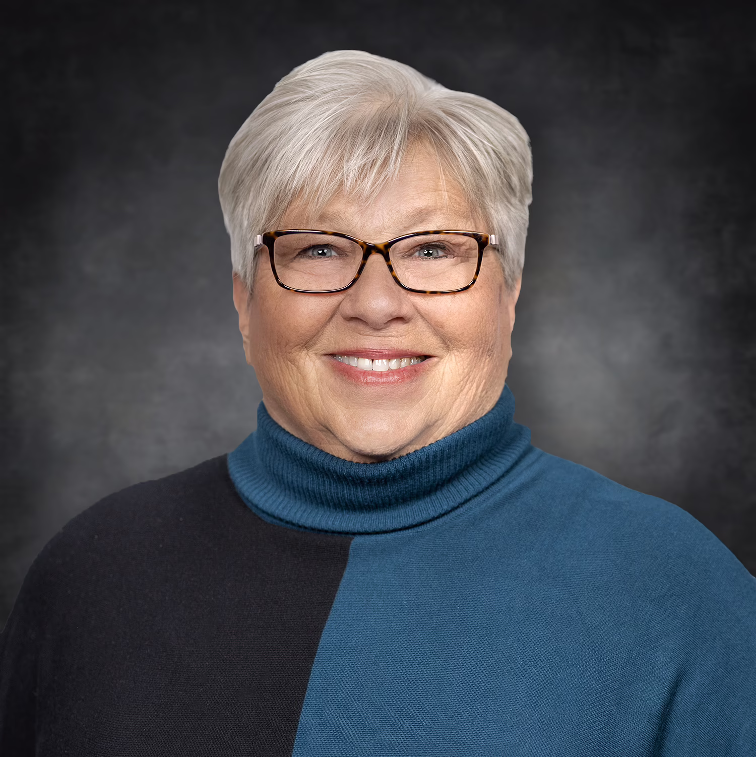 Smiling older woman with short gray hair, glasses, and a teal and black color-block turtleneck sweater poses in front of a dark, softly blurred background.