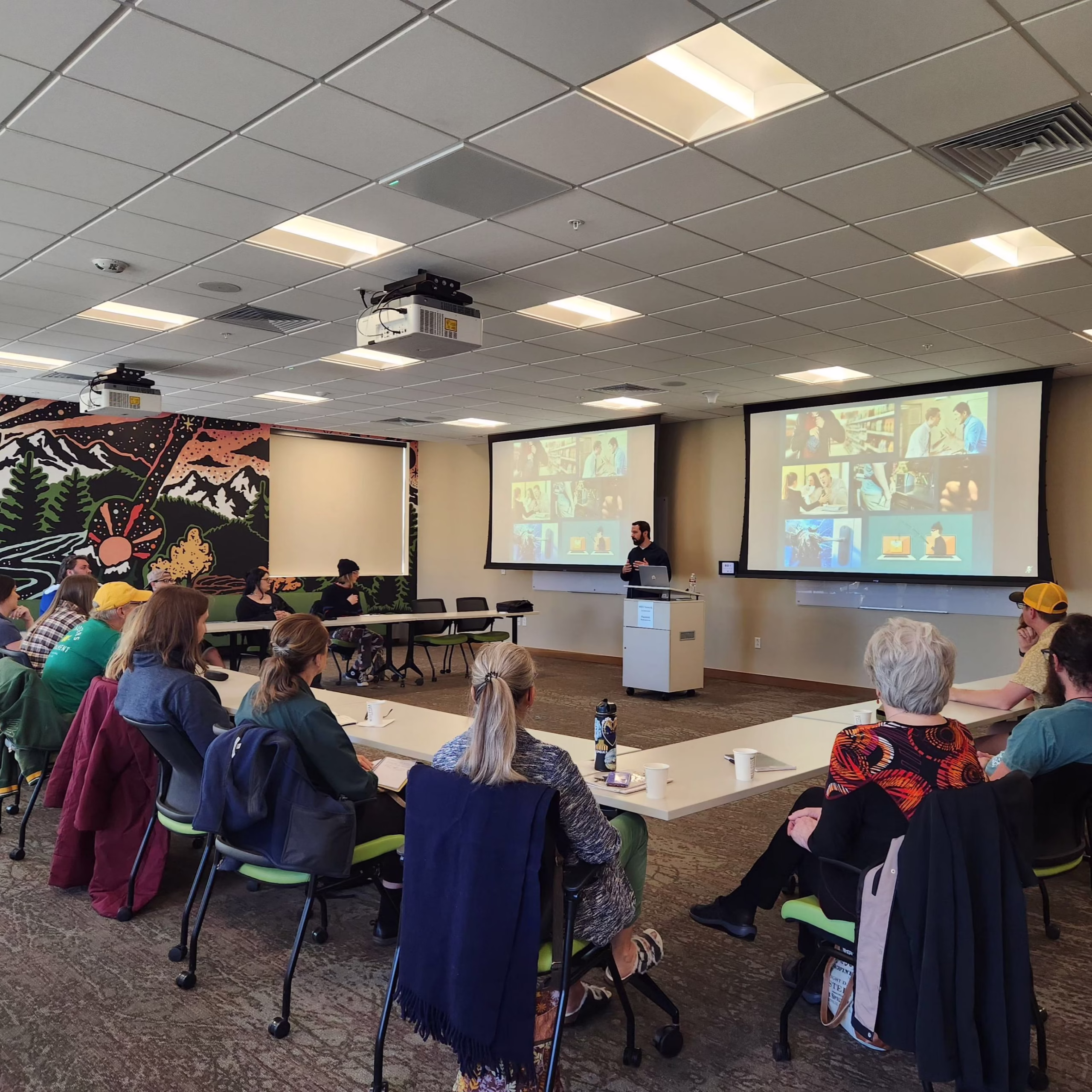 A group of people sit around tables in a conference room, facing a presenter who is speaking at a podium. Two projection screens behind the presenter display slides with images of people.