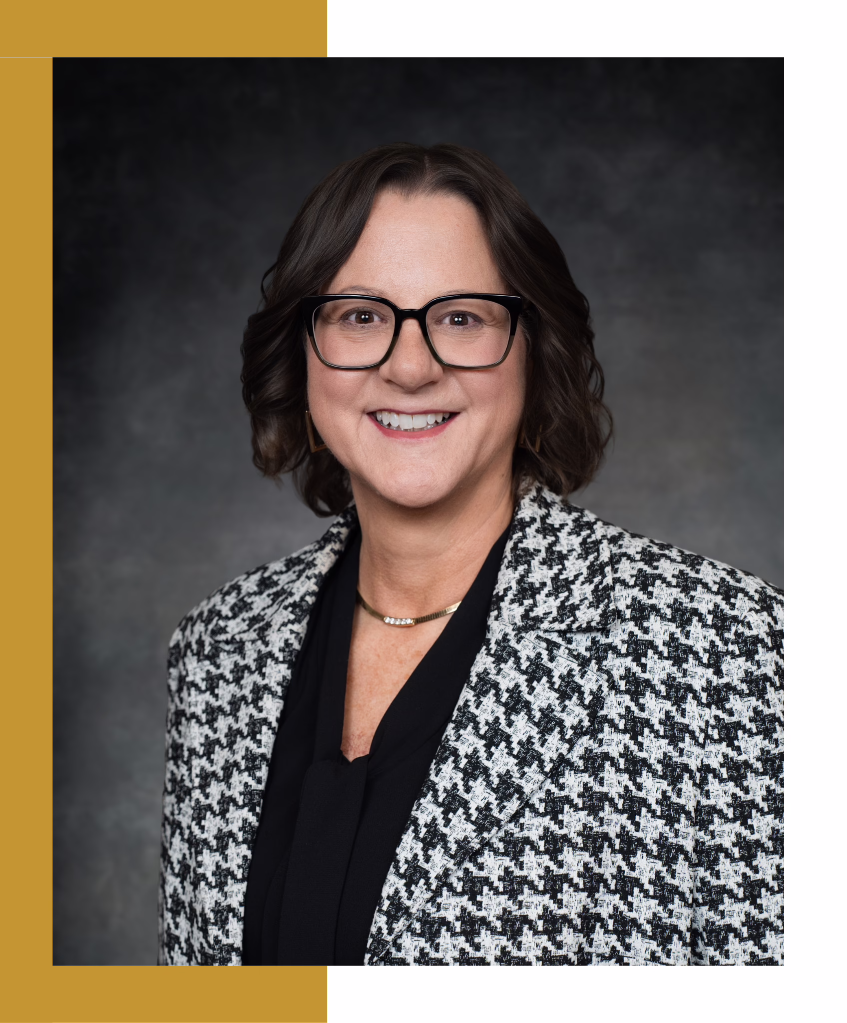 A woman with short brown hair and glasses smiles at the camera. She is wearing a black top and a black-and-white houndstooth blazer, with a dark background and a gold and white border framing the photo.