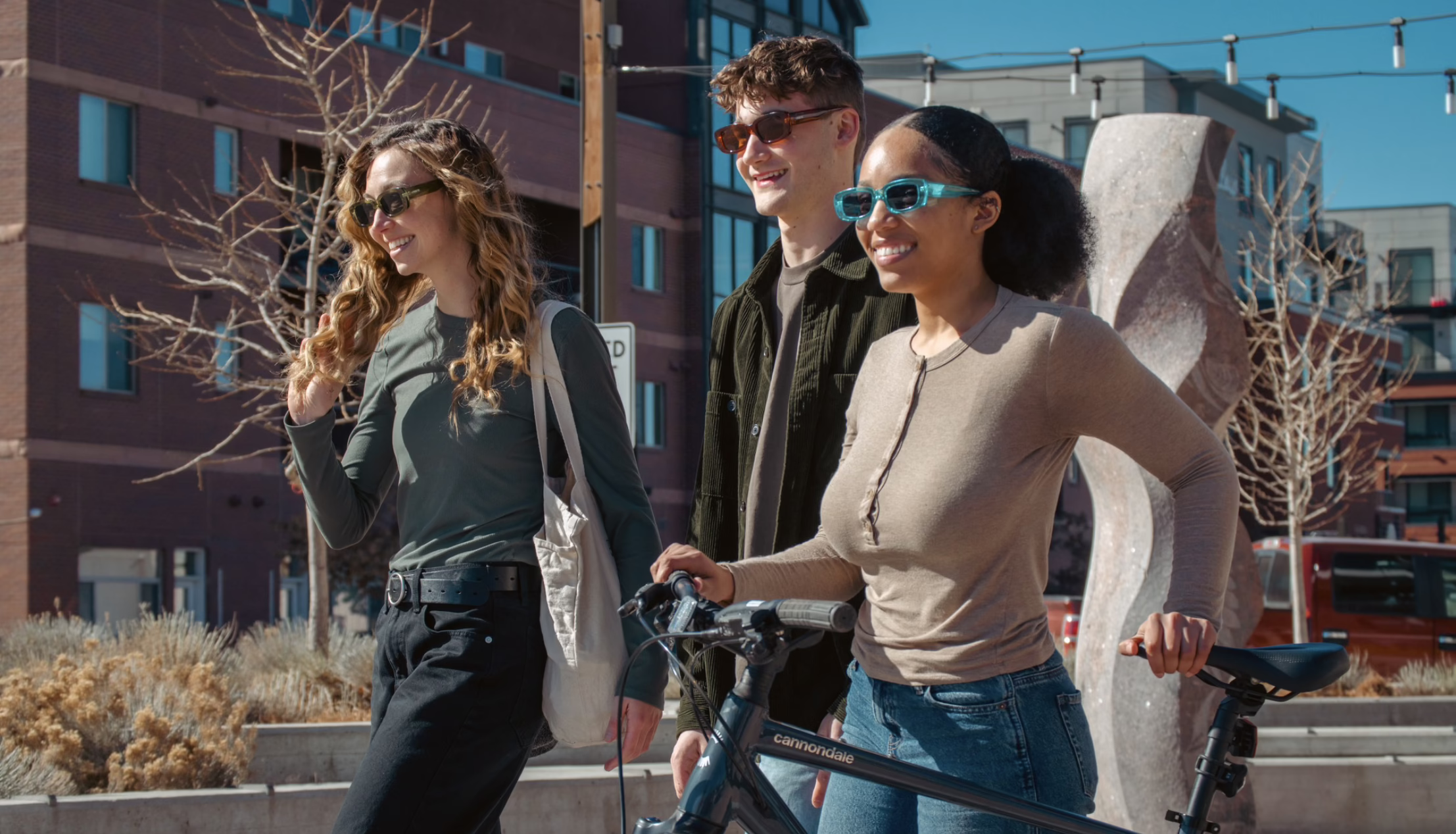 Three smiling young adults wearing sunglasses walk together outdoors in a city setting; one person pushes a bicycle as they enjoy a sunny day near modern buildings and public art.