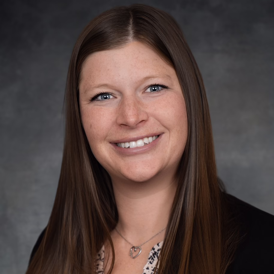 A woman with long straight brown hair, wearing a black blazer, a light patterned blouse, and a heart-shaped necklace, smiling in front of a dark, neutral background.