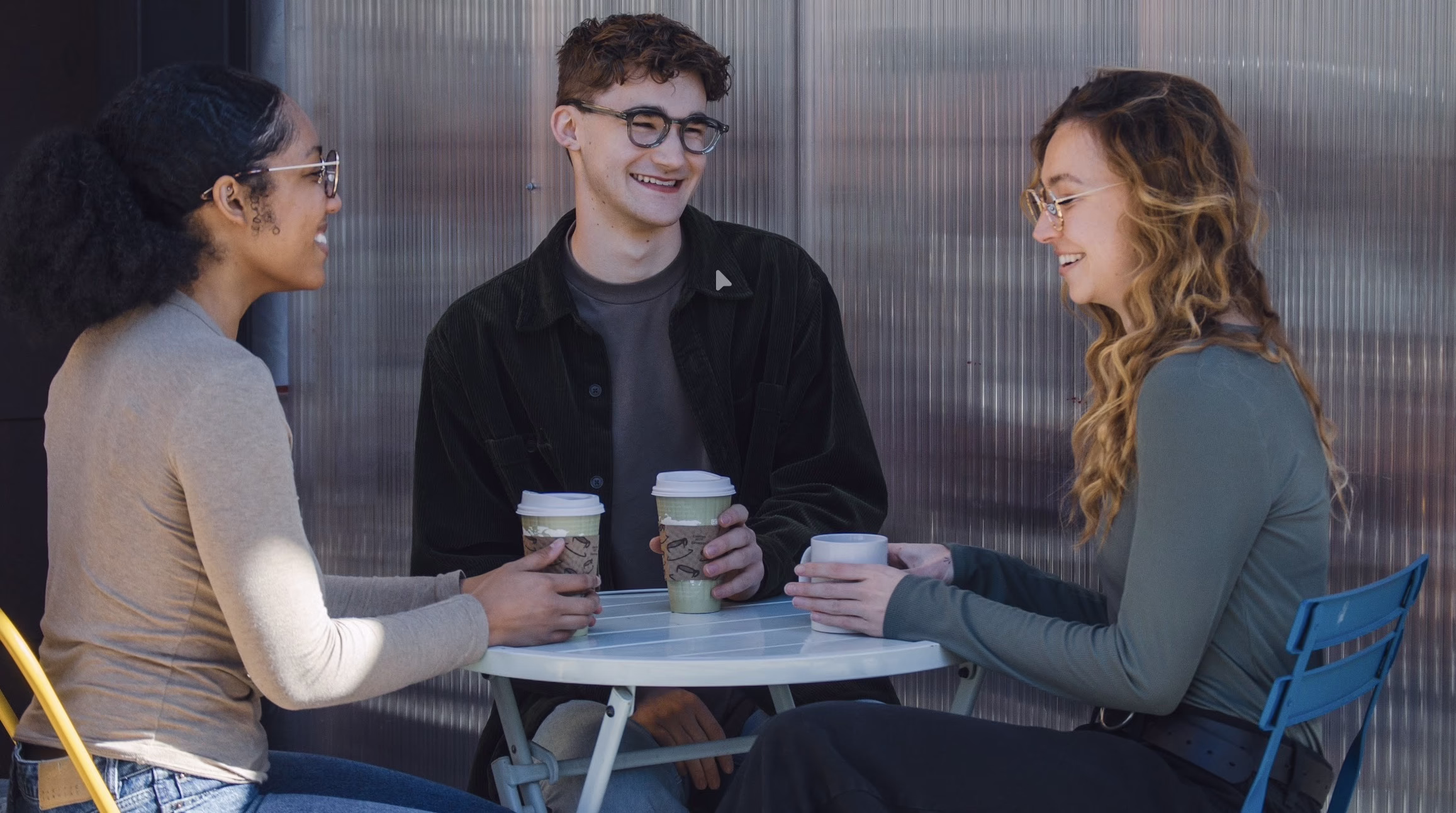 Three young adults sit around a small outdoor table, smiling and talking. Two have takeaway coffee cups, and one has a ceramic mug. They appear relaxed and are enjoying a casual conversation.