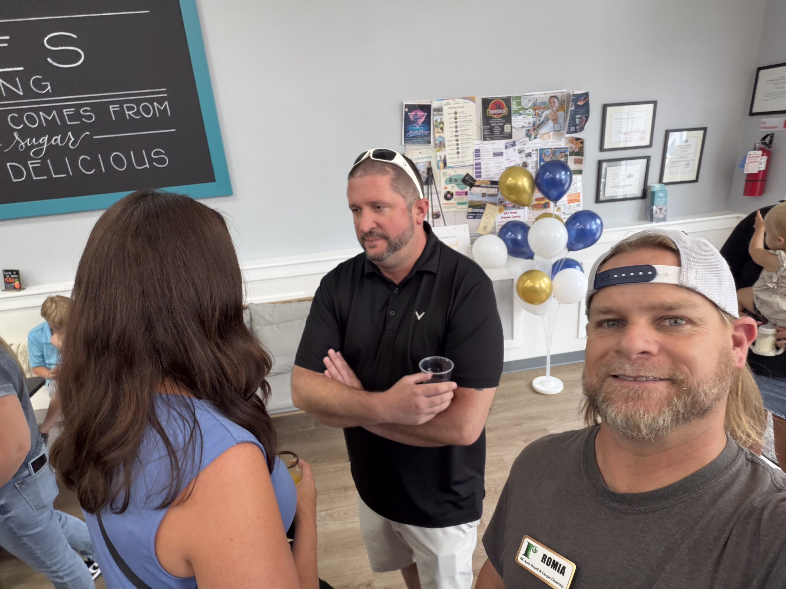 A man with a name tag takes a selfie at an indoor gathering. Two people are talking behind him. There are balloons, a bulletin board, and people in the background. The setting appears casual and friendly.