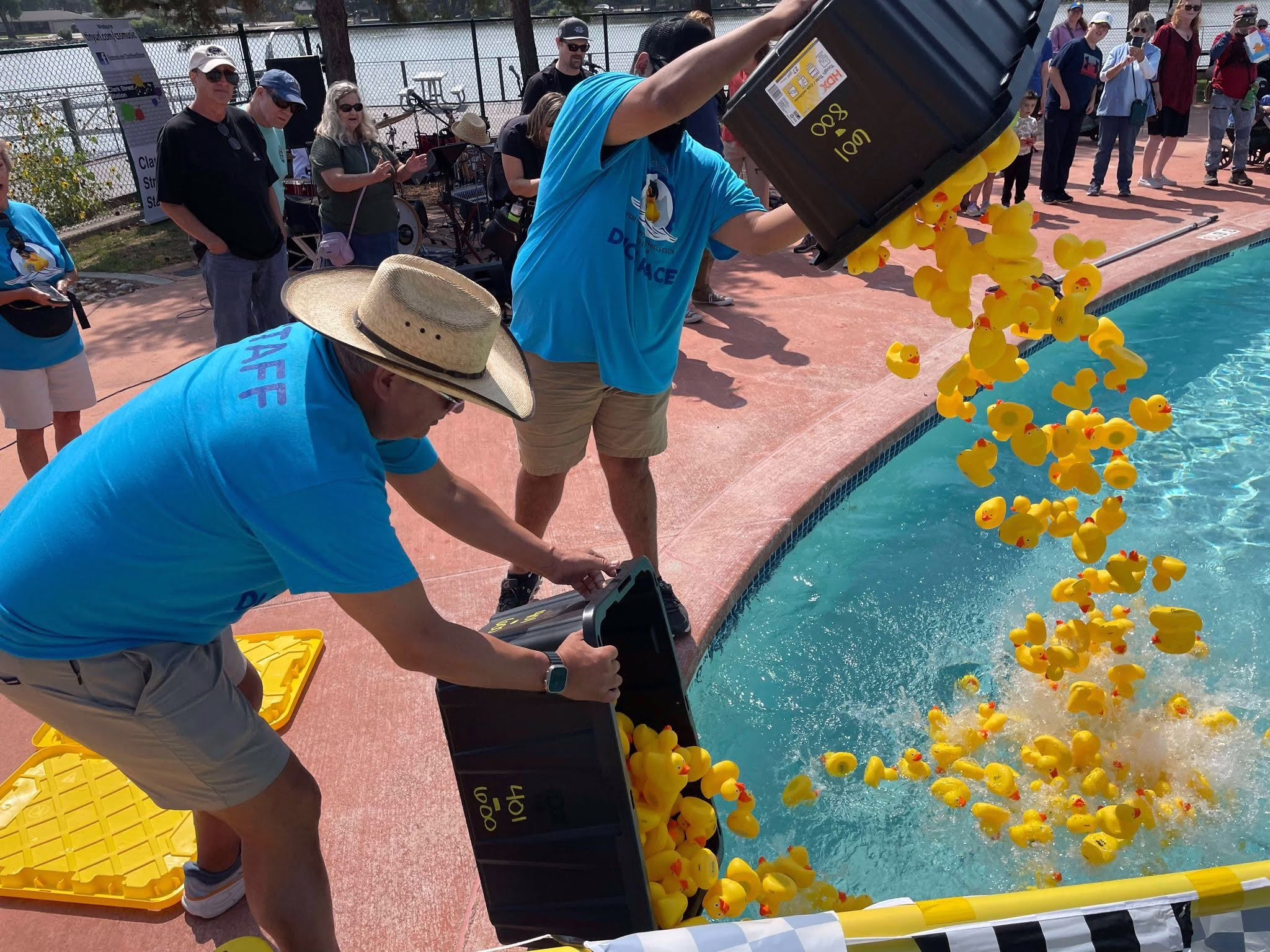 Two people in blue shirts and hats dump large bins of yellow rubber ducks into a pool during an outdoor event, while a crowd watches in the background near a waterfront.