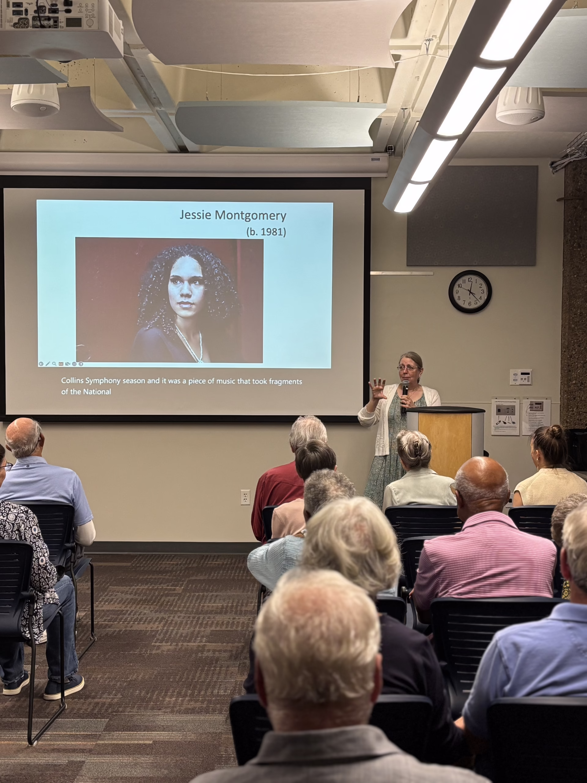 A woman stands at a podium addressing an audience in a classroom. A projected slide behind her shows a photo of Jessie Montgomery, with her name and birth year, and some text about her music.