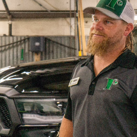 A man with a beard wearing a green cap and a black shirt with a logo stands indoors near a shiny black vehicle, looking to the side and smiling.