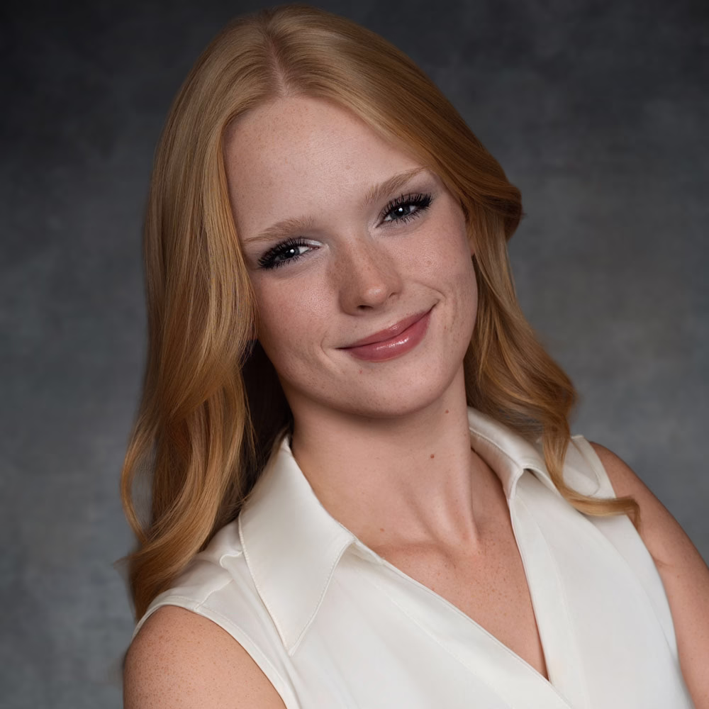 A woman with long red hair and fair skin smiles softly. She wears a sleeveless white blouse and poses in front of a dark, blurred background.