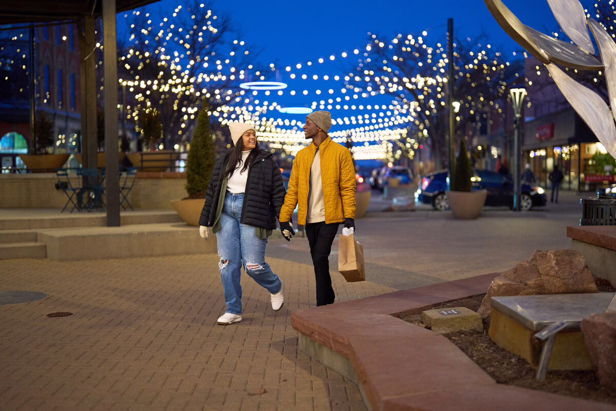 A couple walks hand-in-hand down a pedestrian street decorated with overhead string lights at twilight.